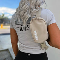 Woman wearing a WW Sahara color crossbody belted bag pictured from back. WEWORKIN BRAND logo imprinted in white and grey ink on the front right lower edge. Smooth matte polyester material, zippered main compartment, color-matched metal zipper pulls on a molded zipper, color-matched interior pocket, with adjustable waist strap.