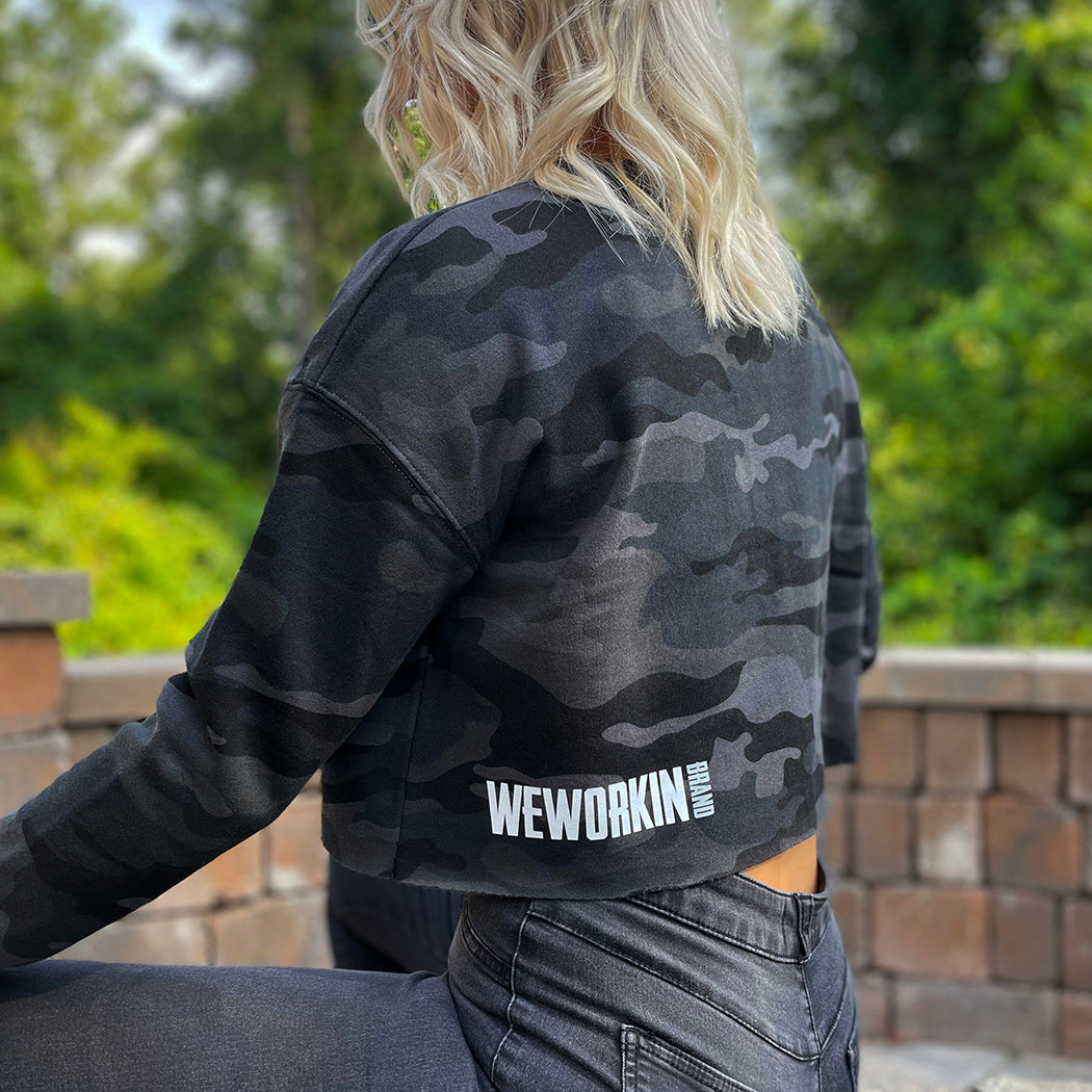 Woman pictured from Back-left side wearing a We Workin women's lightweight cropped crew pullover in Black Camo(back print only, no front print). "WEWORKIN BRAND" block logo is printed on the back, bottom left hem area in white and grey ink. Imprint approx 5.5" wide.