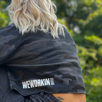 Woman pictured from Back-left side wearing a We Workin women's lightweight cropped crew pullover in Black Camo(back print only, no front print). "WEWORKIN BRAND" block logo is printed on the back, bottom left hem area in white and grey ink. Imprint approx 5.5" wide.