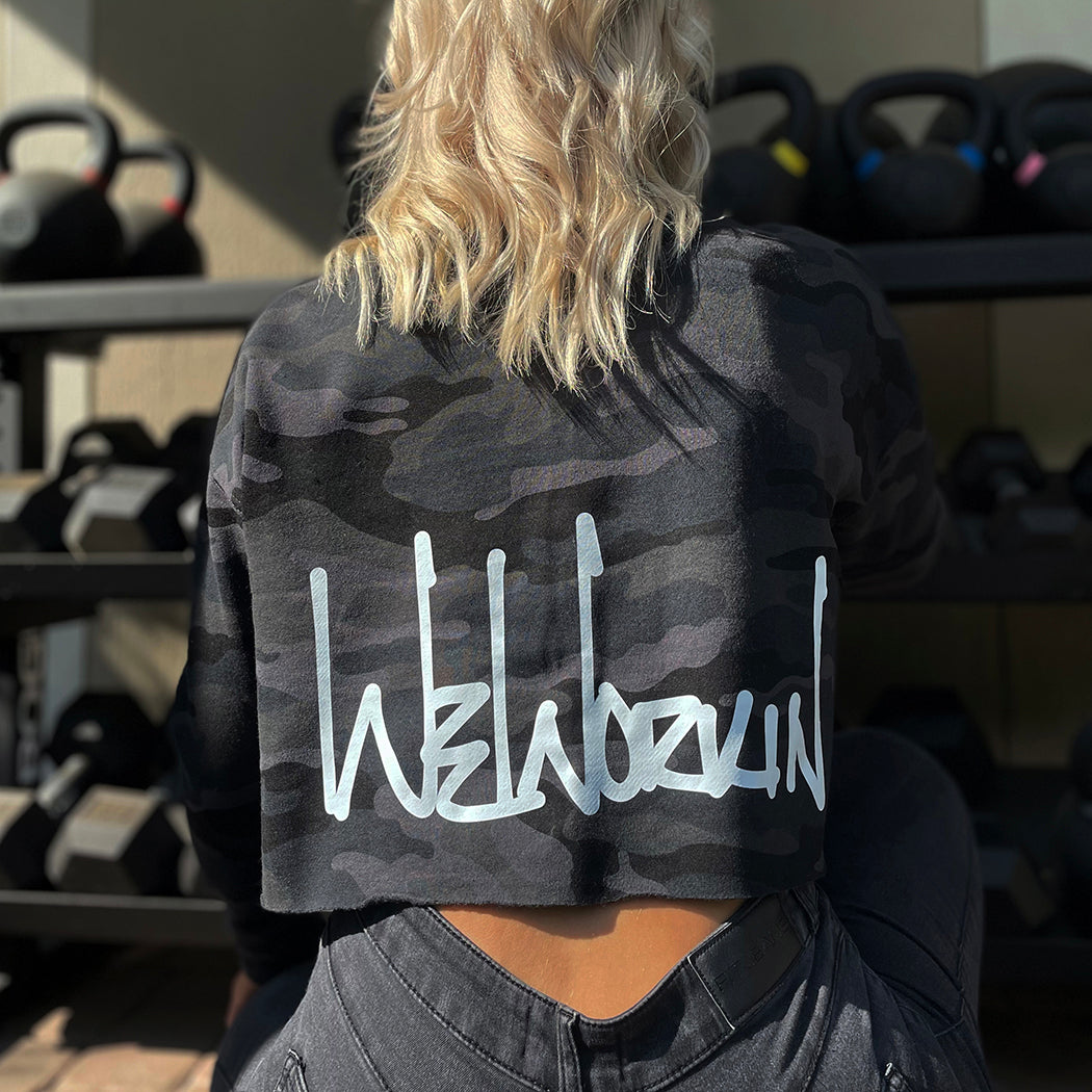 Woman pictured from back wearing a We Workin women's lightweight cropped crew pullover in Black Camo (back print only, no front print). WEWORKIN Script logo is printed large across the back, bottom hem area in white ink. Imprint approx 13" wide.