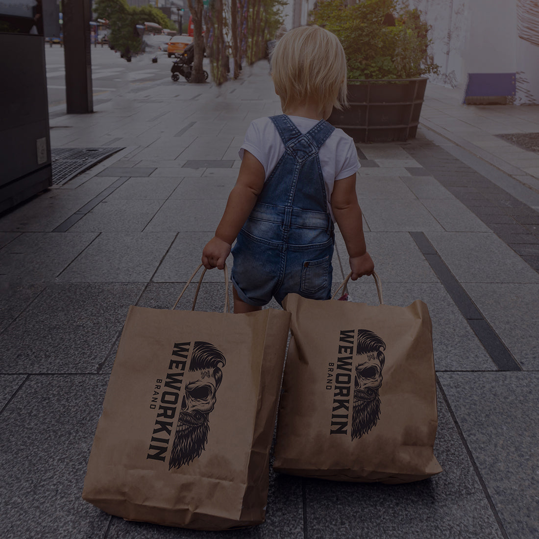Toddler in jean overalls dragging 2 large stuffed WEWORKIN shopping bags down a city sidewalk