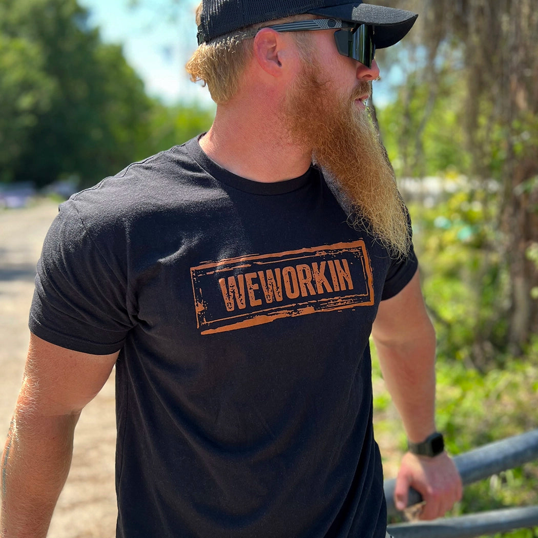 Man pictured outdoors from front, wearing a We Workin black graphic tee, boldly screenprinted with our original design "WEWORKIN Dirt Stamp" graphic, on the full front chest, in brown ink.