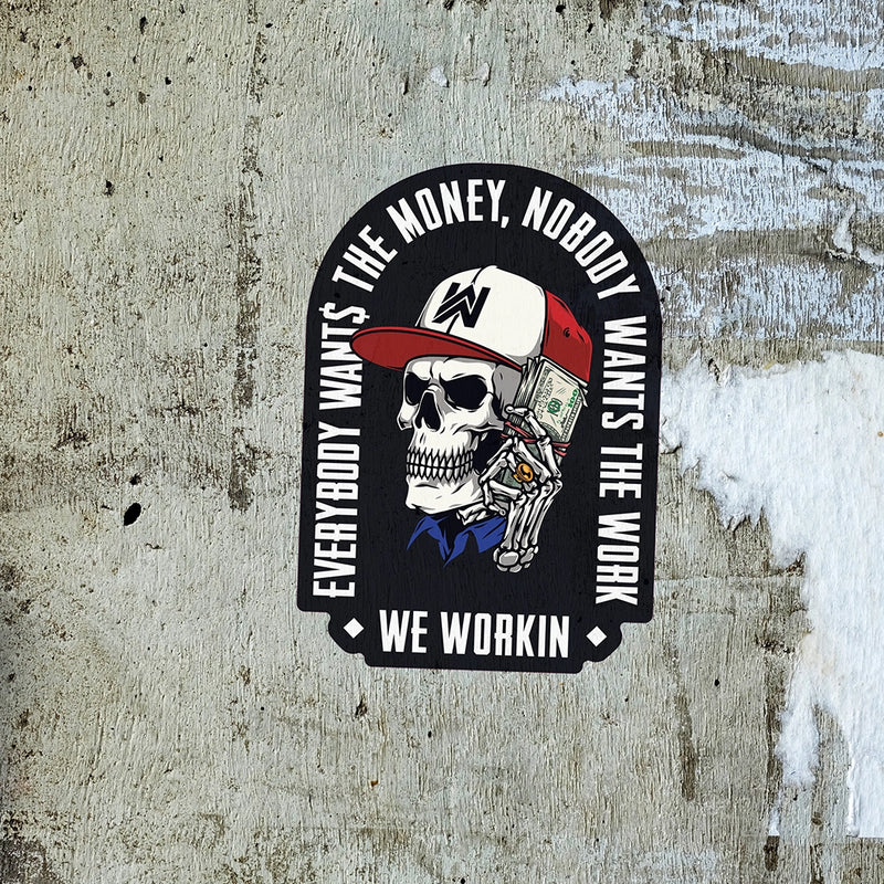 EVERYBODY WANT$ THE MONEY, NOBODY WANTS THE WORK. WE WORKIN—Die-cut sticker. "EWTM" tagline and WE WORKIN text, surrounding a Skull wearing a hat and holding a money bundle to his ear. Skull graphic in color, text in white. EWTM Arch design on black sticker, placed on a grungey concrete wall.