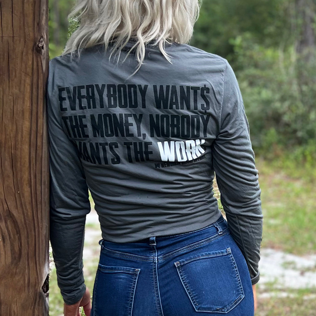 Woman pictured outdoors from the back, wearing a WE WORKIN lightweight long sleeve tee in Smoke Grey color. The back is printed with the tagline "Everybody Wants the Money, Nobody Wants the WORK." in black ink, except the word WORK is in white ink.