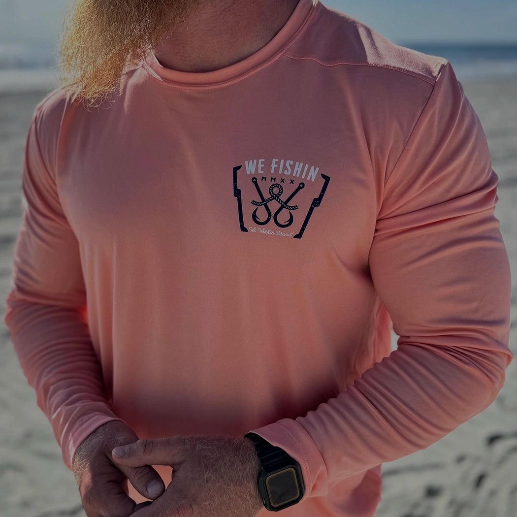 FISH Collection. Man pictured from front outdoors at the ocean, wearing a WW FISH Long Sleeve ultra lightweight shirt in coral, screen printed with our newest design, our WE FISHIN and fishhooks/line design on the left chest "pocket" area.