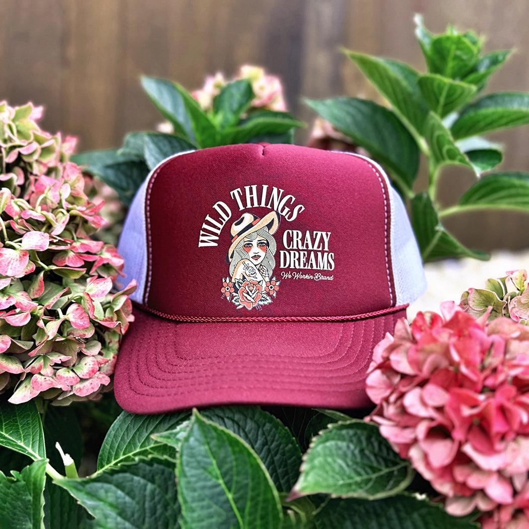 A WeWorkin Foam Trucker Snapback Hat in a Maroon/White colorway, in a floral background, pictured from the front. The design shows “WILD THINGS—CRAZY DREAMS” text surrounding a full-color stylized tatted-woman image on the full front foam panel. We Workin Brand text in cursive in the bottom right of design.