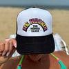 Woman wearing a WeWorkin Foam Trucker Snapback Hat in Black/White/Black colorway, pictured outdoors from front. Design shows WE WORKIN text in Neon Vibe colors arched over THE WORKIN CLASS stacked text. MM and XX are on the left and right respectively.