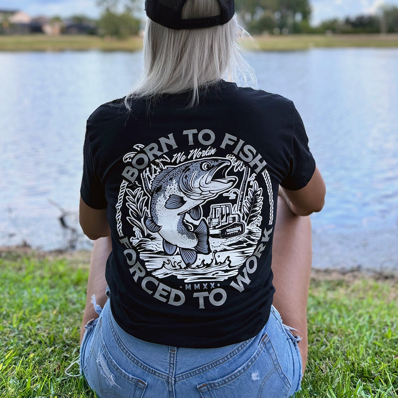 Woman wearing a WW black tee, shown from back, sitting along a water bank. BORN TO FISH—FORCED TO WORK text encircles a graphic of Fish jumping out of water and bulldozer on land in the background. Printed in White and Grey inks. Graphic is printed large on the centerback.