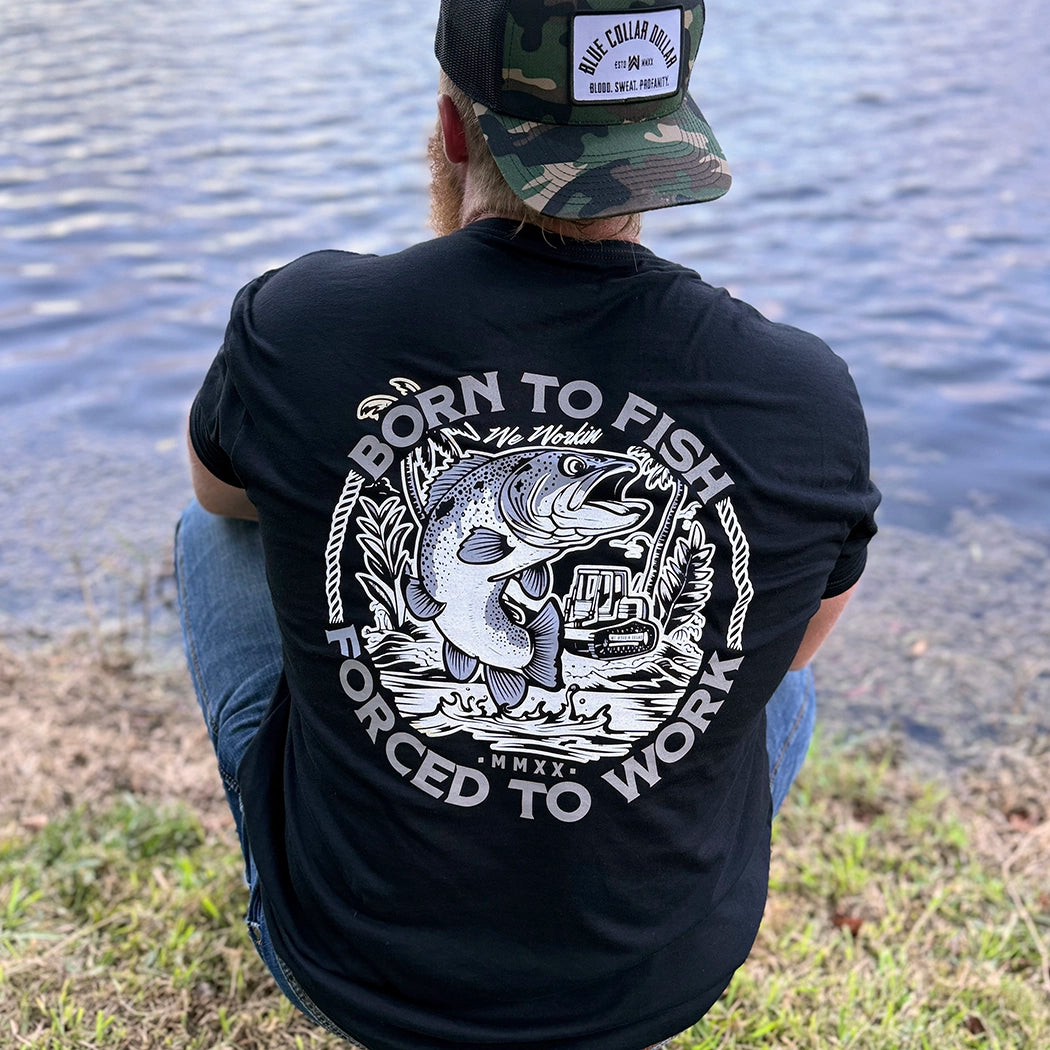Man wearing a WW black tee, shown from back, sitting along a water bank. BORN TO FISH—FORCED TO WORK text encircles a graphic of Fish jumping out of water and bulldozer on land in the background. Printed in White and Grey inks. Graphic is printed large on the centerback.