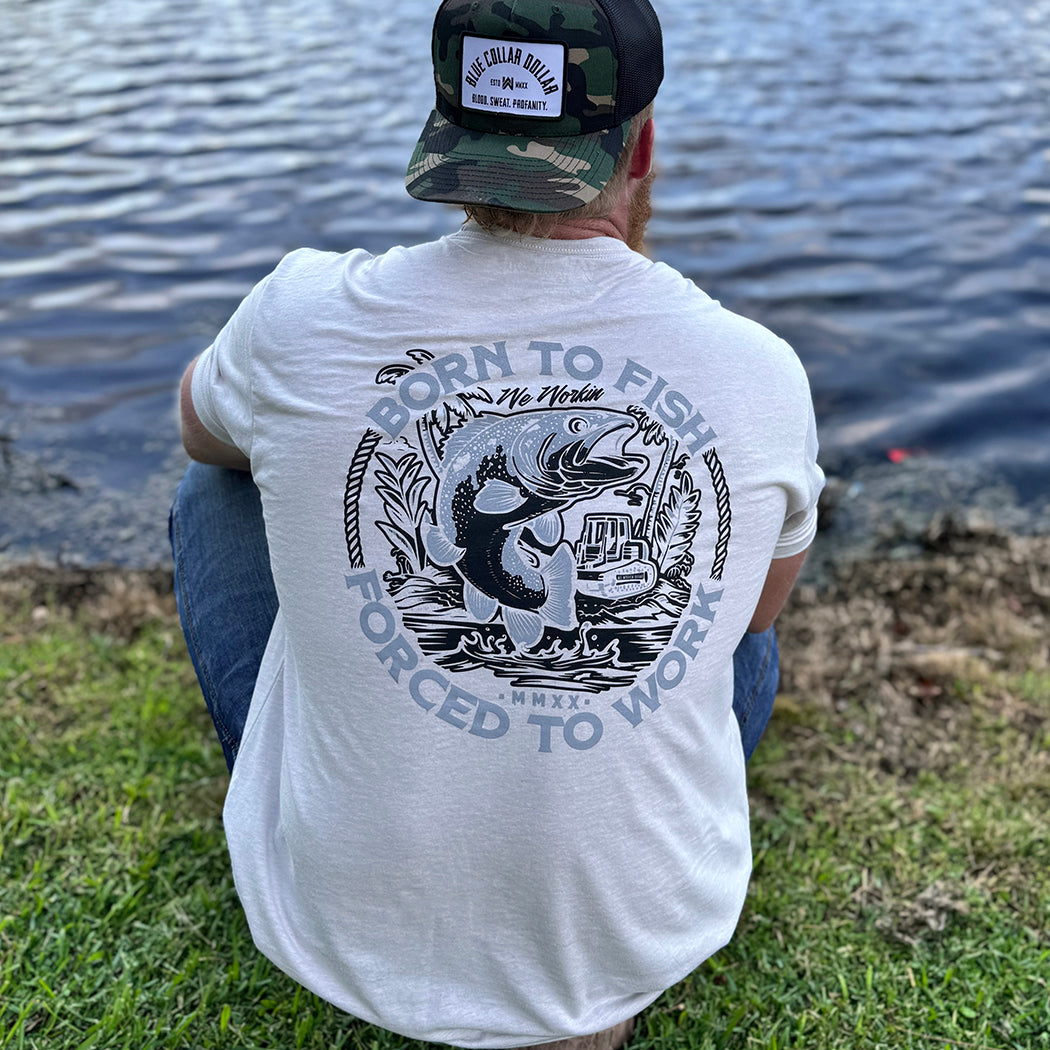 Man wearing a WW sand color tee, shown from back, sitting along a water bank. BORN TO FISH—FORCED TO WORK text encircles a graphic of Fish jumping out of water and bulldozer on land in the background. Printed in Black and Grey inks. Graphic is printed large on the centerback.