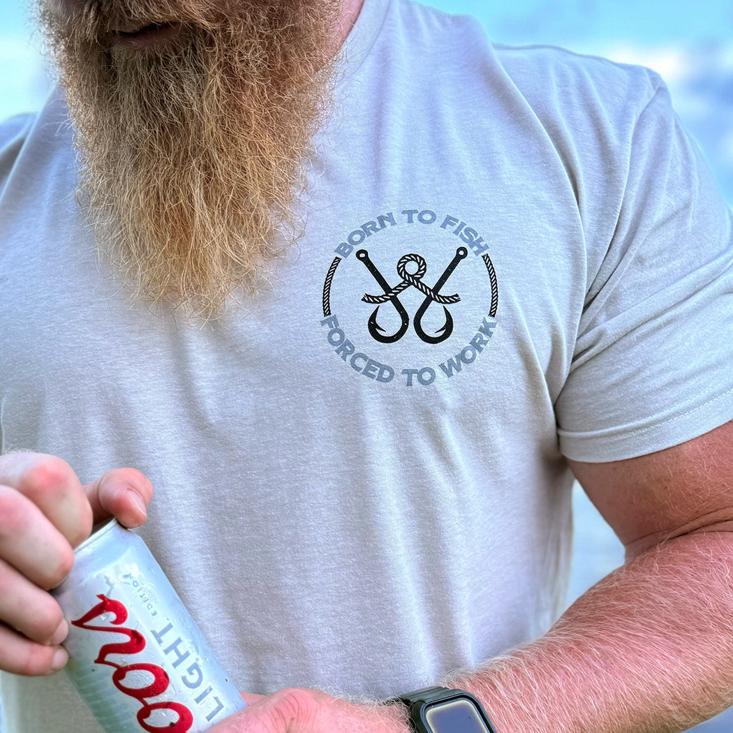 Man wearing a WW sand color tee, shown from front. BORN TO FISH—FORCED TO WORK text encircles fishing hooks and line (printed on the left chest "pocket" area.) Printed in Black and Grey inks.