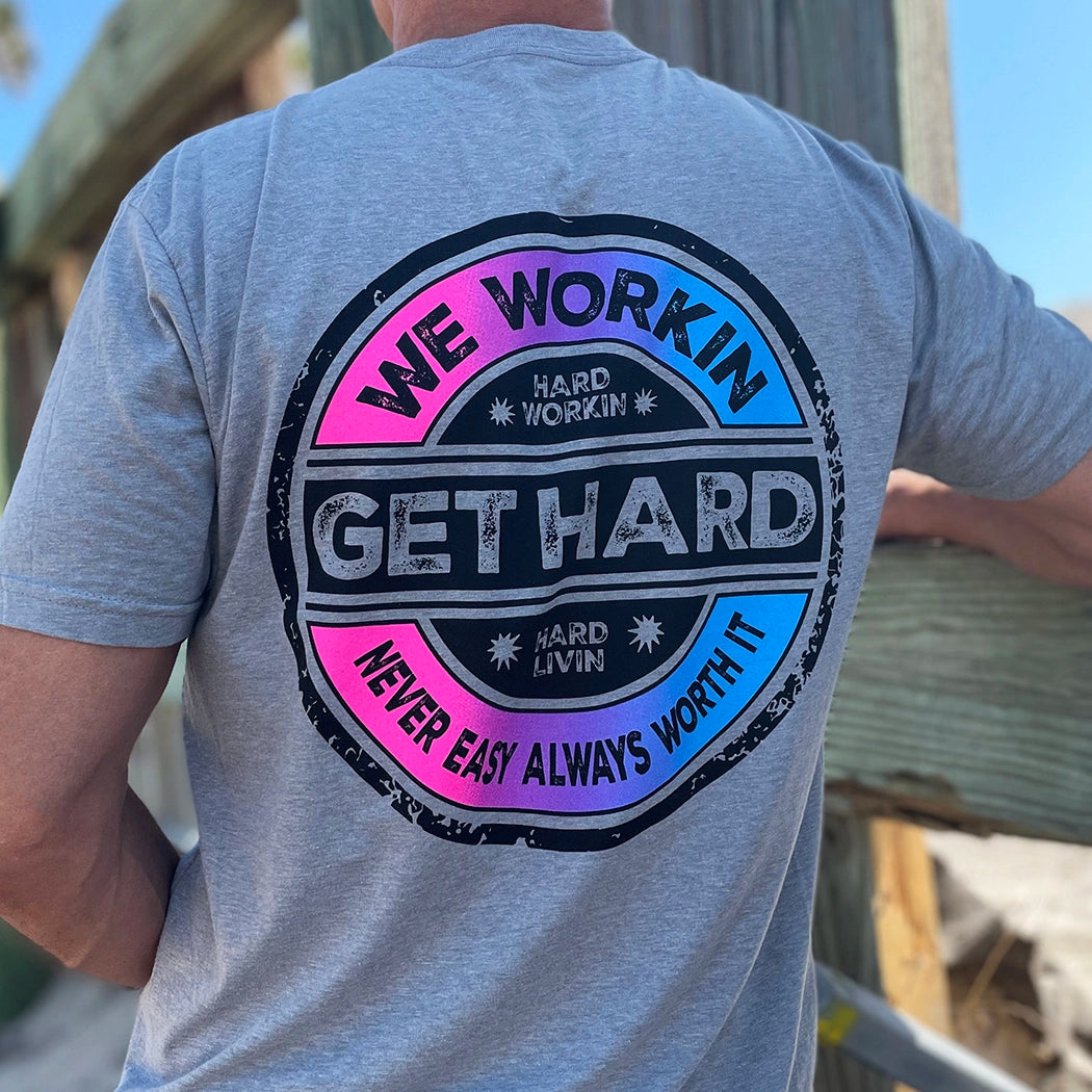 Man from back, wearing a WW heather grey tee. 3-color Distressed circular Neon Pink and Bright Blue faded design, "GET HARD" main text in center of layout, "WE WORKIN" and "Never Easy Always Worth It" on the top and bottom of circular design, and finally "Hard Workin" and "Hard Livin" in center top/bottom—printed large in the center/upper back.
