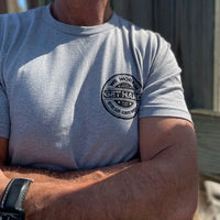 Man from front, wearing a WW dark heather grey tee. 1-color black, distressed circular design, "GET HARD" main text in center of layout, "WE WORKIN" and "Never Easy Always Worth It" on the top and bottom of circular design, and finally "Hard Workin" and "Hard Livin" in center top/bottom—printed small in the left chest "pocket" area.