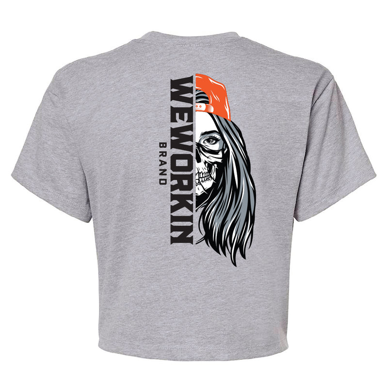 Back of a We Workin Women's short-sleeve cropped tee in heather grey—with a large imprint of our "WEWORKIN BRAND vertical text and Half Skull Woman with Hat" design in Black/White/Grey (hat graphic highlighted in Orange ink.)