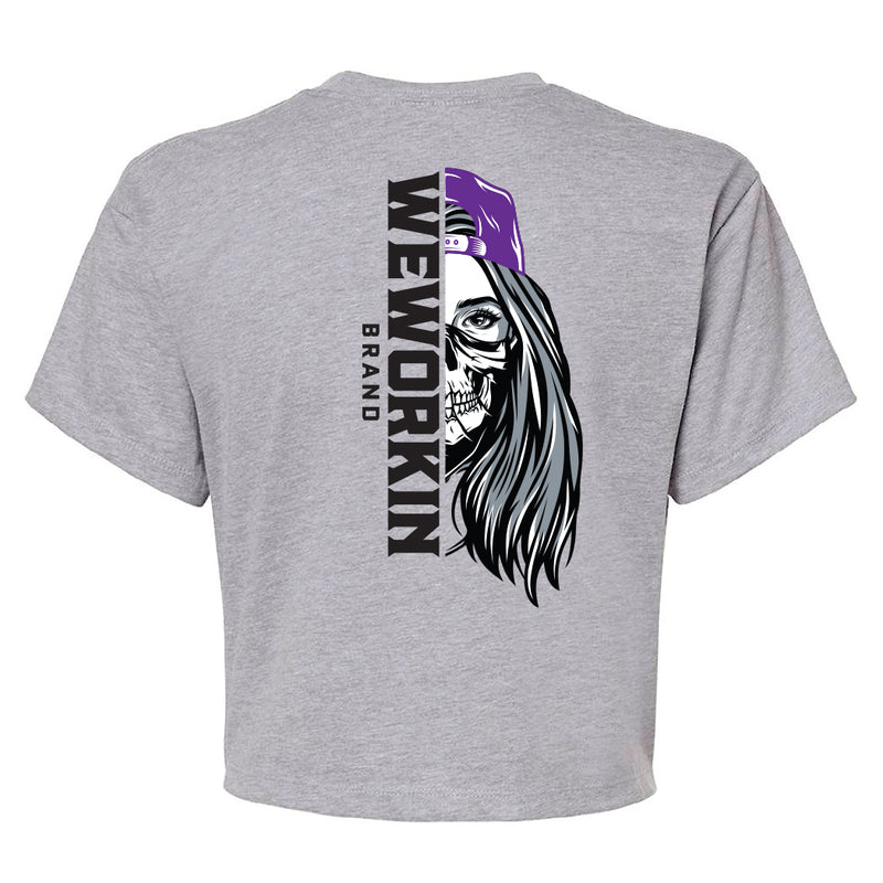 Back of a We Workin Women's short-sleeve cropped tee in heather grey—with a large imprint of our "WEWORKIN BRAND vertical text and Half Skull Woman with Hat" design in Black/White/Grey (hat graphic highlighted in Purple ink.)
