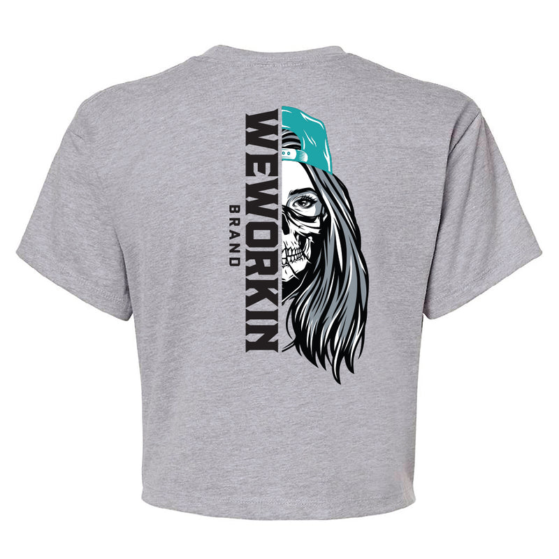 Back of a We Workin Women's short-sleeve cropped tee in heather grey—with a large imprint of our "WEWORKIN BRAND vertical text and Half Skull Woman with Hat" design in Black/White/Grey (hat graphic highlighted in Teal ink.)