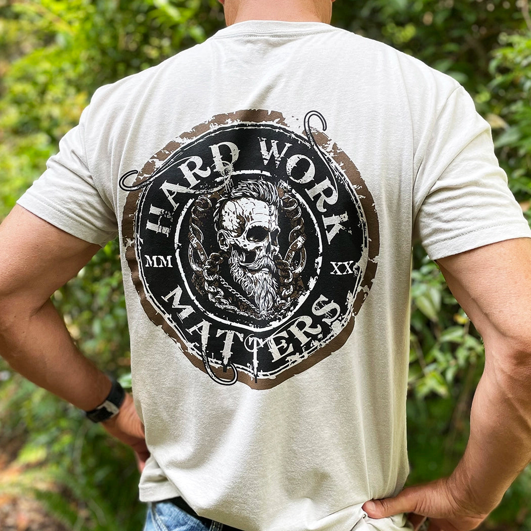 Man wearing a WW Sand color Graphic Tee (pictured from back), imprinted with "HARD WORK MATTERS" circular text and a rugged skull image in Viking-style design in center of circular image, on full back width in Black, Brown and White inks. (MMXX Roman numerals on the left/right of design printed small.)