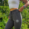 Woman pictured from front-left side wearing the We Workin women's joggers in Heathered Charcoal Grey. "NOT FOR THE WEAK" white text encircling the WW icon logo in grey, subtly printed just below the front left pocket area. [Design approx 2.25" wide.]