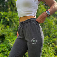 Woman pictured from front-left side wearing the We Workin women's joggers in Heathered Charcoal Grey. "NOT FOR THE WEAK" white text encircling the WW icon logo in grey, subtly printed just below the front left pocket area. [Design approx 2.25" wide.]