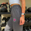 Woman pictured from front-left side wearing the We Workin women's joggers in Heathered Charcoal Grey. "NOT FOR THE WEAK" white text encircling the WW icon logo in bright pink, subtly printed just below the front left pocket area. [Design approx 2.25" wide.]