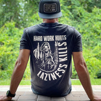 Man pictured from back wearing a We Workin graphic tee in a Charcoal Grey color. Athletic/Slimmer fit, short sleeve shirt, full back imprinted with a cloaked grim-reaper in chains flippin the bird graphic—and large text HARD WORK HURTS. LAZINESS KILLS (printed in white ink.) Also wearing a WW Flat bill Patch hat in Grey/black.