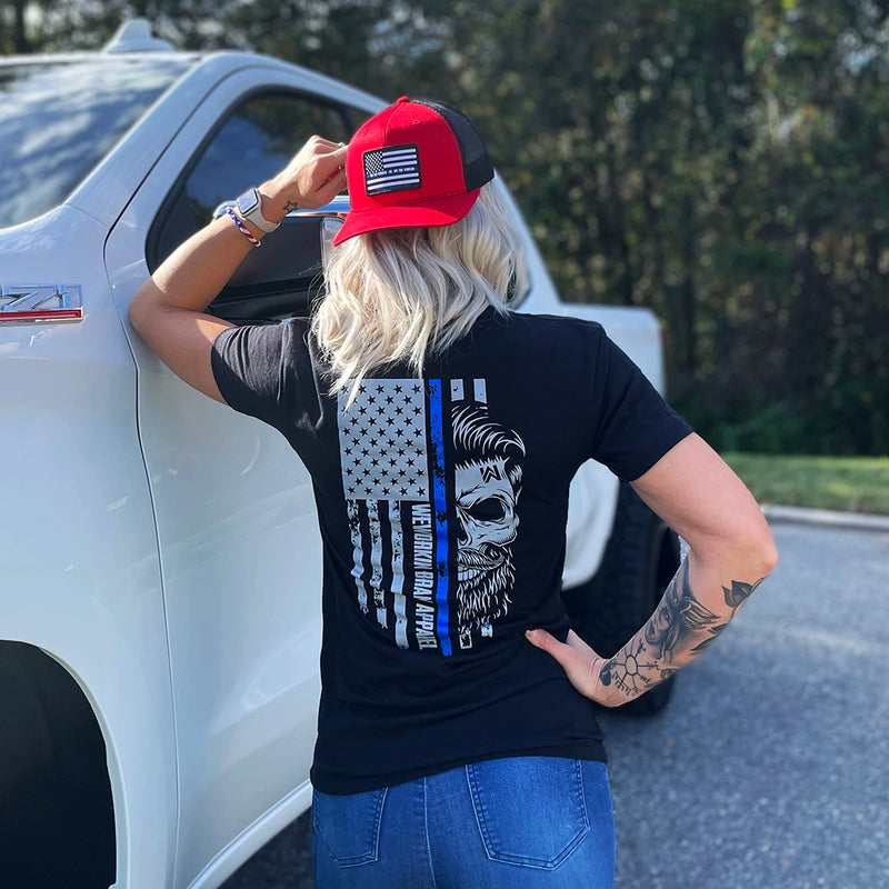 Woman outdoors, pictured from back, wearing a We Workin black, short sleeve men's tee with the LEO Flag Thin Blue Line WWB design, printed large on the back. Also wearing a Flat bill 511 red-black hat, with LEO-Flag patch.