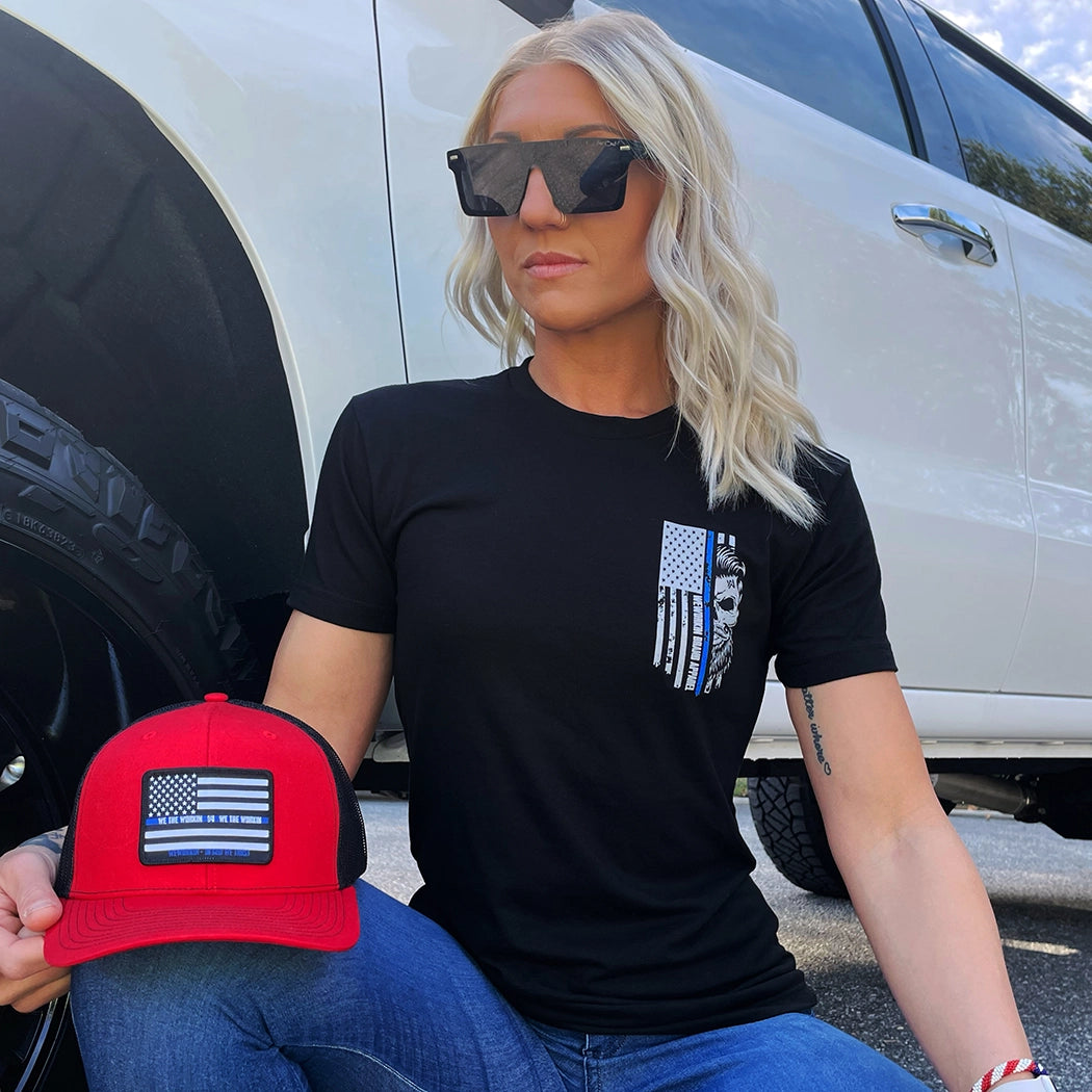 Woman outdoors, pictured from front, wearing a We Workin black, short sleeve men's tee with the LEO Flag Thin Blue Line WWB design, printed small on the front left chest "pocket" area. Also showing a Flat bill 511 red-black hat, with LEO-Flag patch.