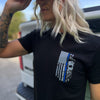 Woman outdoors, pictured from front close up, wearing a We Workin black, short sleeve men's tee with the LEO Flag Thin Blue Line WWB design, printed small on the front left chest "pocket" area.