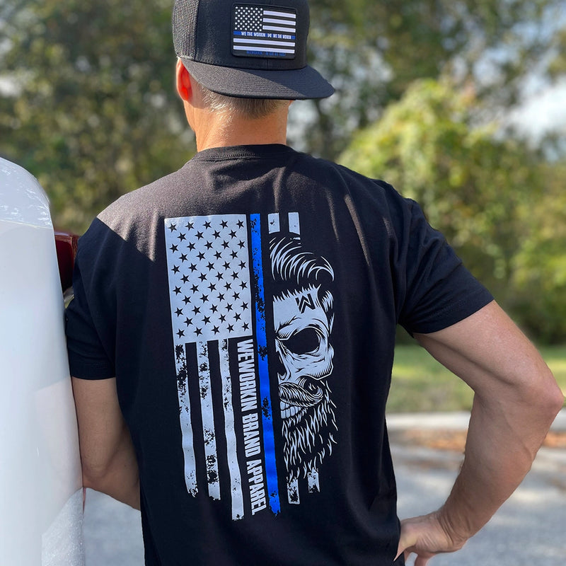 Male outdoors, pictured from back, wearing a We Workin black, short sleeve tee with the LEO Flag Thin Blue Line WWB design, printed large on the back. Also wearing a Flat bill 511 all black LEO-Flag patch hat.