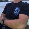 Male outdoors, pictured from front close up, wearing a We Workin black, short sleeve tee with the LEO Flag Thin Blue Line WWB design, printed small on the left front chest "pocket" area. 