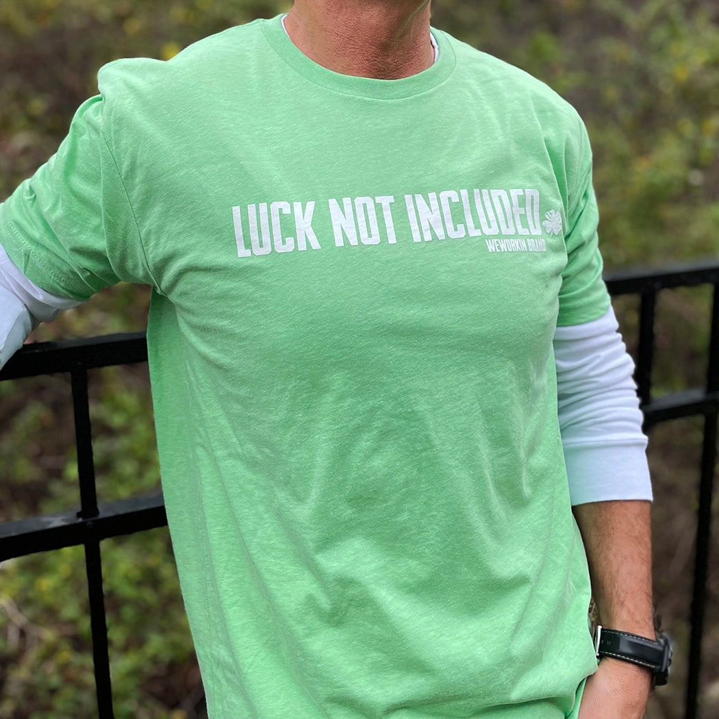 Man pictured from the front wearing a Green WeWorkin short sleeve tee. Printed large across front of chest, in white ink, LUCK NOT INCLUDED with a 4-leaf clover at the end (WEWORKIN BRAND  is underneath main text on right side, small font). 