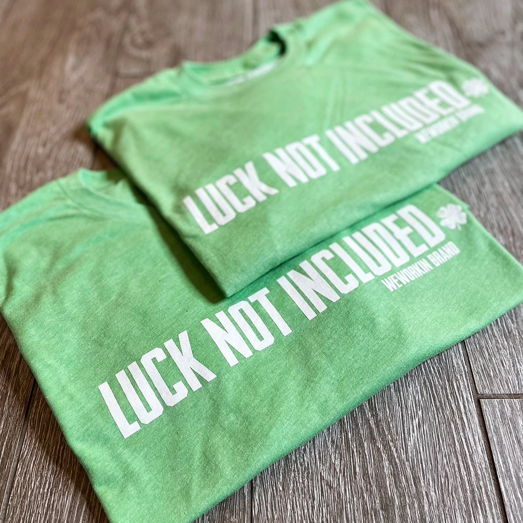 Pic of two folded Green color WeWorkin short sleeve tee on tile background. Printed large across front of chest, in white ink, LUCK NOT INCLUDED with a 4-leaf clover at the end (WEWORKIN BRAND is underneath main text on right side, small font). 