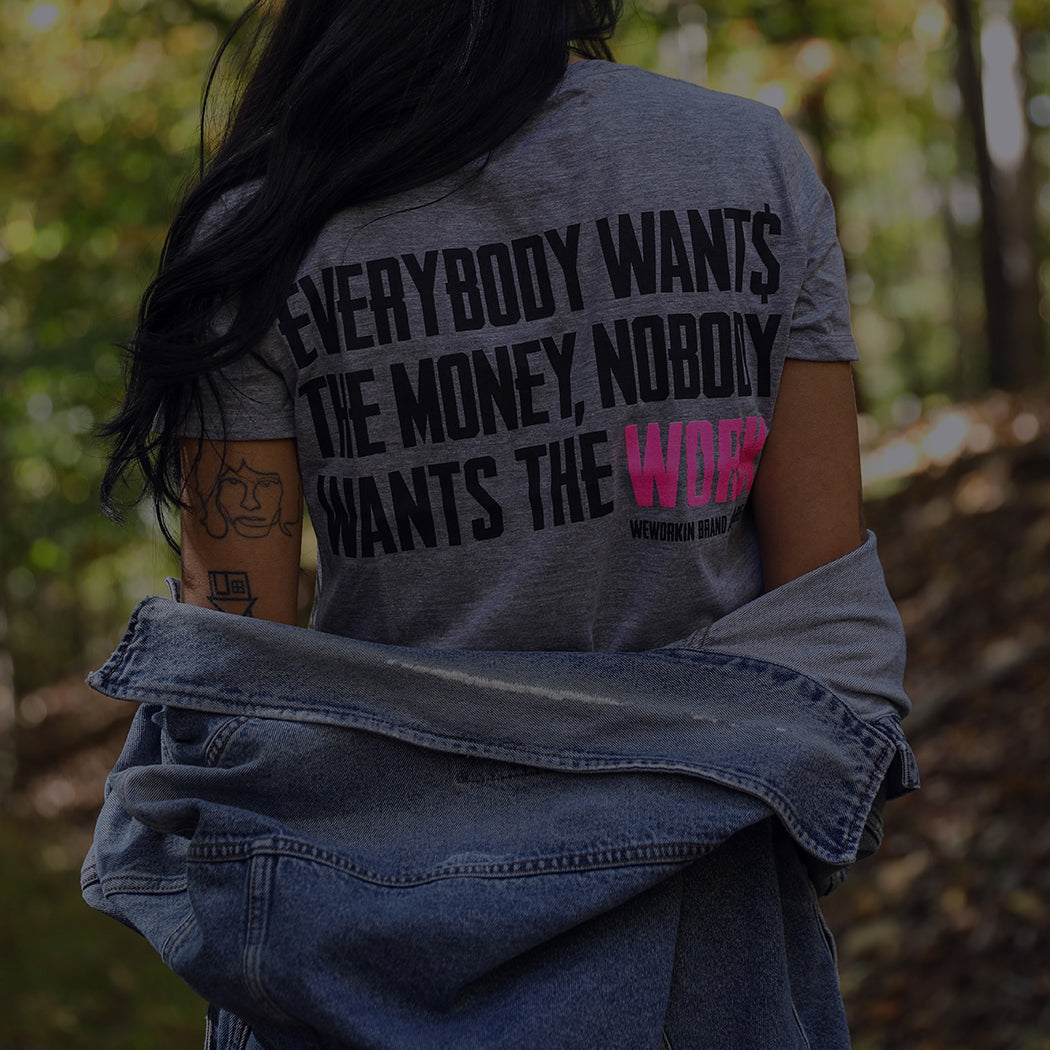 Women's Collection. Woman is wearing a Grey tee with the tagline "Everybody Want$ the Money. Nobody Wants the WORK." printed on the back in white/pink inks.