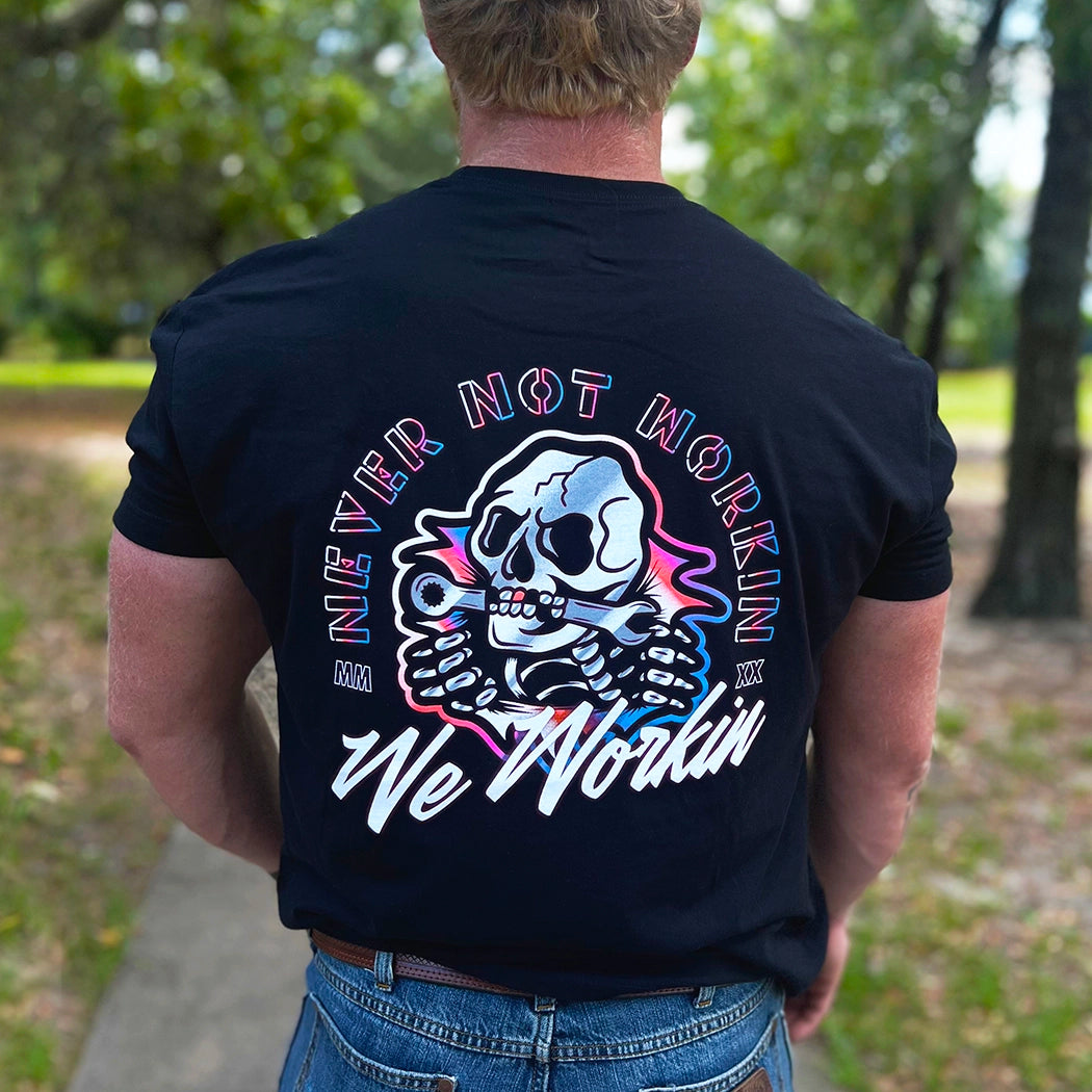 Man wearing a WW black tee, shown from back. "NEVER NOT WORKIN" text arching around the top and sides of a Neon Skull "bustin out" as center image. WE WORKIN is along the bottom of the graphic in script. Graphic is printed large on the centerback. Printed in white and multiple Neon inks.