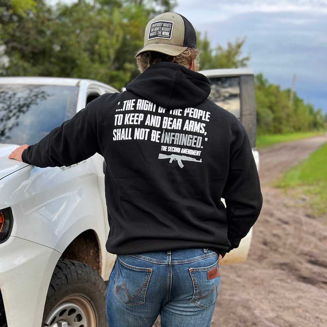 Man pictured from back, outdoors, wearing a WW PRO-2A black hooded pullover sweatshirt. "...The right of the people to keep and bear arms, shall not be infringed." design is printed on the back in white ink, with grey ink highlighting "infringed." and an AR. Banded cuffs and waist. 