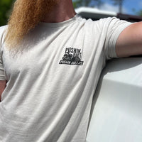 Man pictured from front wearing a WW Desert Sand color Graphic Tee. "PUSHIN DIRT. PUSHIN DOLLAR$" bulldozer graphic printed in black, in left "pocket" area.