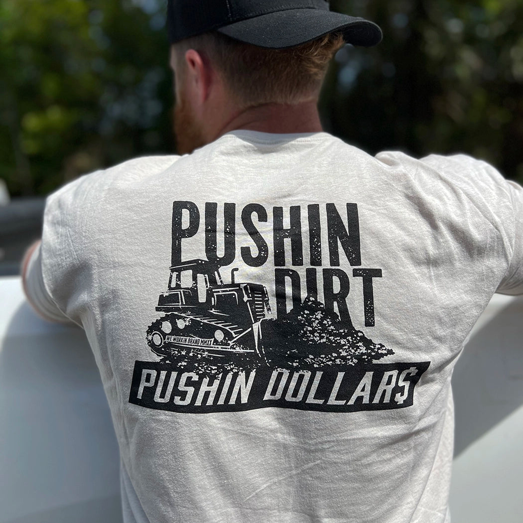 Man pictured from back wearing a WW Desert Sand color Graphic Tee. "PUSHIN DIRT. PUSHIN DOLLAR$" bulldozer graphic printed in black, across full upper back area.