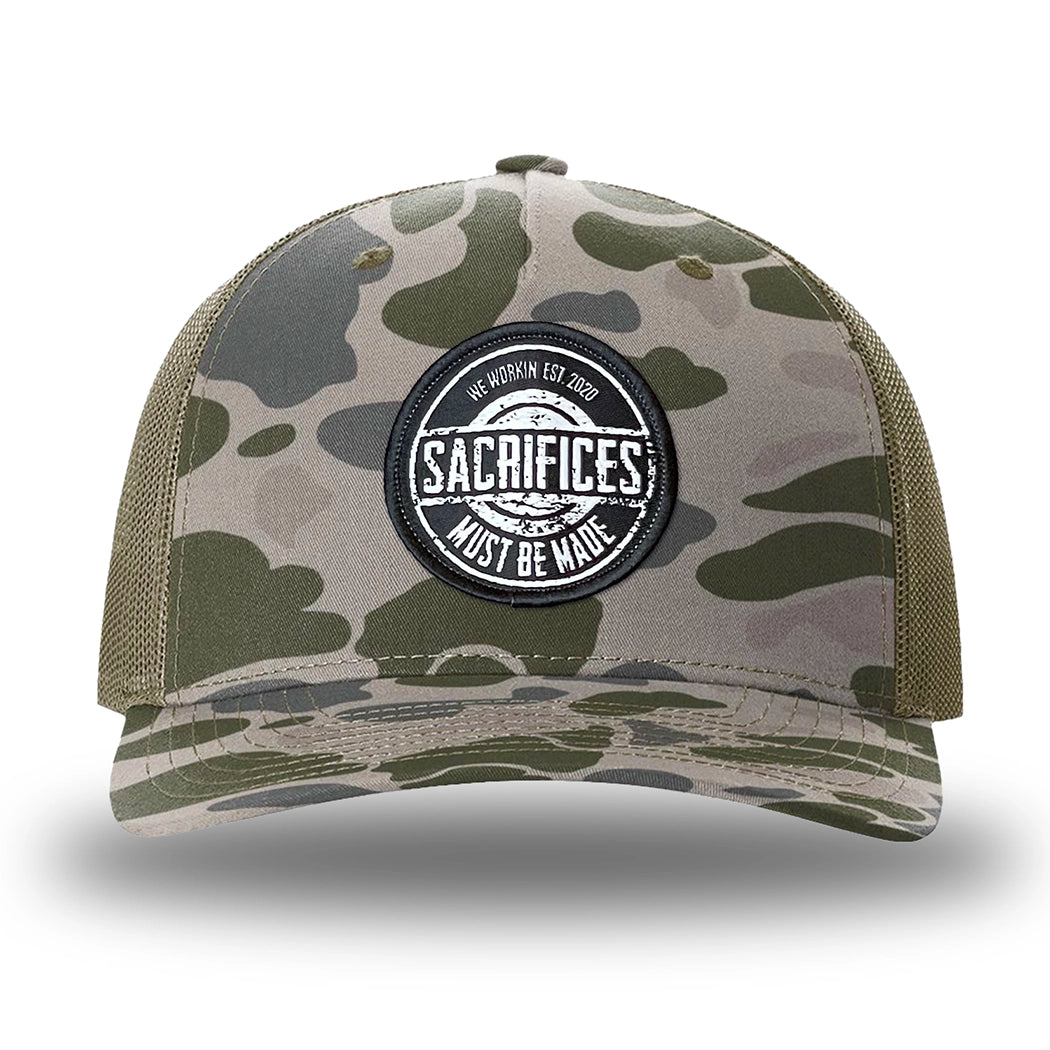 Marsh Duck Camo/Loden two-tone WeWorkin hat—Richardson 112PFP snapback, 5-panel trucker, mesh-back style. WeWorkin "SACRIFICES MUST BE MADE" circular woven patch is centered on the front panel.