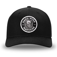 All Black Flex Fit style WeWorkin hat—Woven front with Poly mesh sides and back, Richardson 110 brand (R-Flex trucker). HARD WORK MATTERS woven patch with white merrowed edge, on a black background with HARD WORK MATTERS text encircling a Viking-style skull center graphic with MM XX on the left and right respectively—patch is centered on the front panels.