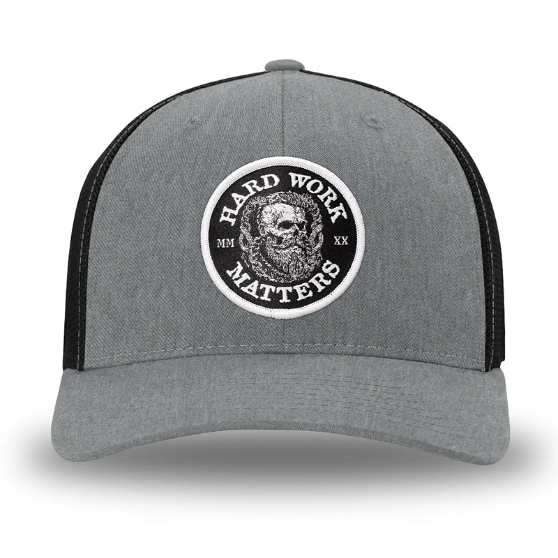 Heather Grey/Black Flex Fit style WeWorkin hat—Woven front with Poly mesh sides and back, Richardson 110 brand (R-Flex trucker). HARD WORK MATTERS woven patch with white merrowed edge, on a black background with HARD WORK MATTERS text encircling a Viking-style skull center graphic with MM XX on the left and right respectively—patch is centered on the front panels.