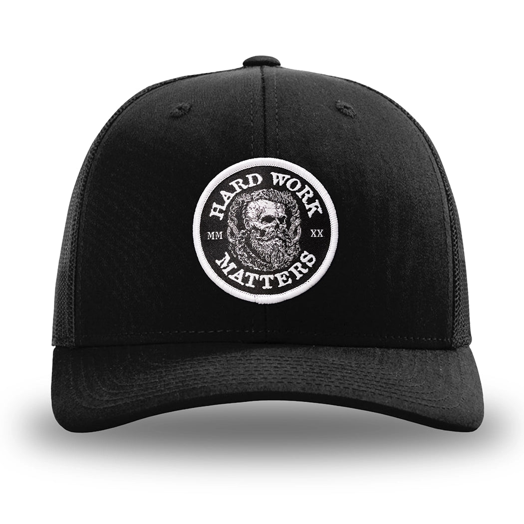 Solid Black WeWorkin hat—Richardson 112 brand snapback, retro trucker classic hat style. HARD WORK MATTERS woven patch with white merrowed edge, on a black background with HARD WORK MATTERS text encircling a Viking-style skull center graphic with MM XX on the left and right respectively—patch is centered on the front panels.