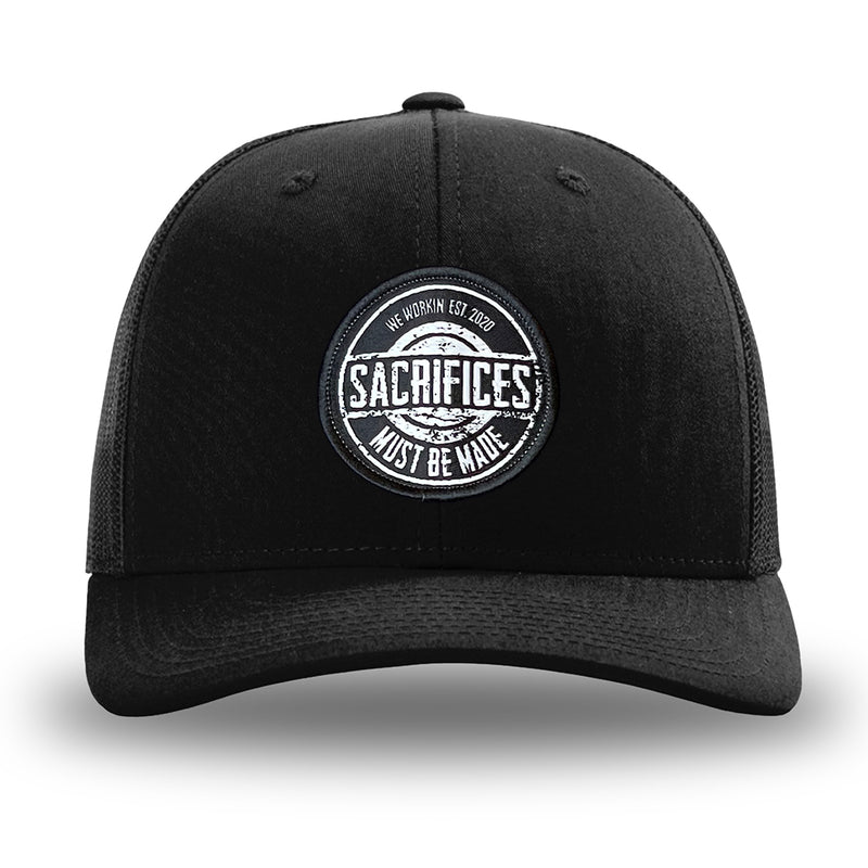 Solid Black WeWorkin hat—Richardson 112 brand snapback, retro trucker classic hat style. WeWorkin "SACRIFICES Must Be Made" circular woven patch, with black/white thread colors and black merrowed edge, is centered on the front panels.