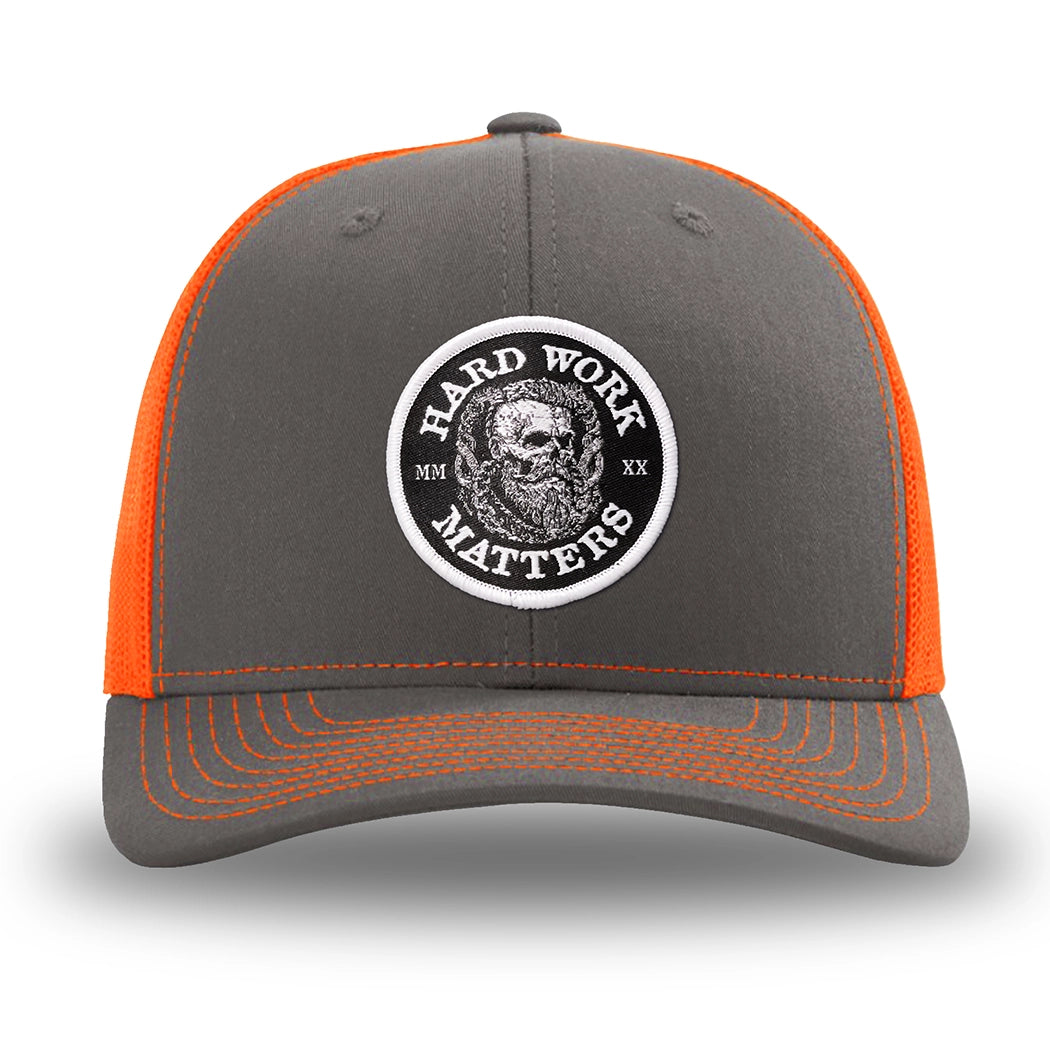 Neon/Safety Orange and Charcoal Grey two-tone WeWorkin hat—Richardson 112 brand snapback, retro trucker classic hat style. HARD WORK MATTERS woven patch with white merrowed edge, on a black background with HARD WORK MATTERS text encircling a Viking-style skull center graphic with MM XX on the left and right respectively—patch is centered on the front panels.