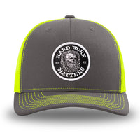 Neon/Safety Yellow and Charcoal Grey two-tone WeWorkin hat—Richardson 112 brand snapback, retro trucker classic hat style. HARD WORK MATTERS woven patch with white merrowed edge, on a black background with HARD WORK MATTERS text encircling a Viking-style skull center graphic with MM XX on the left and right respectively—patch is centered on the front panels.