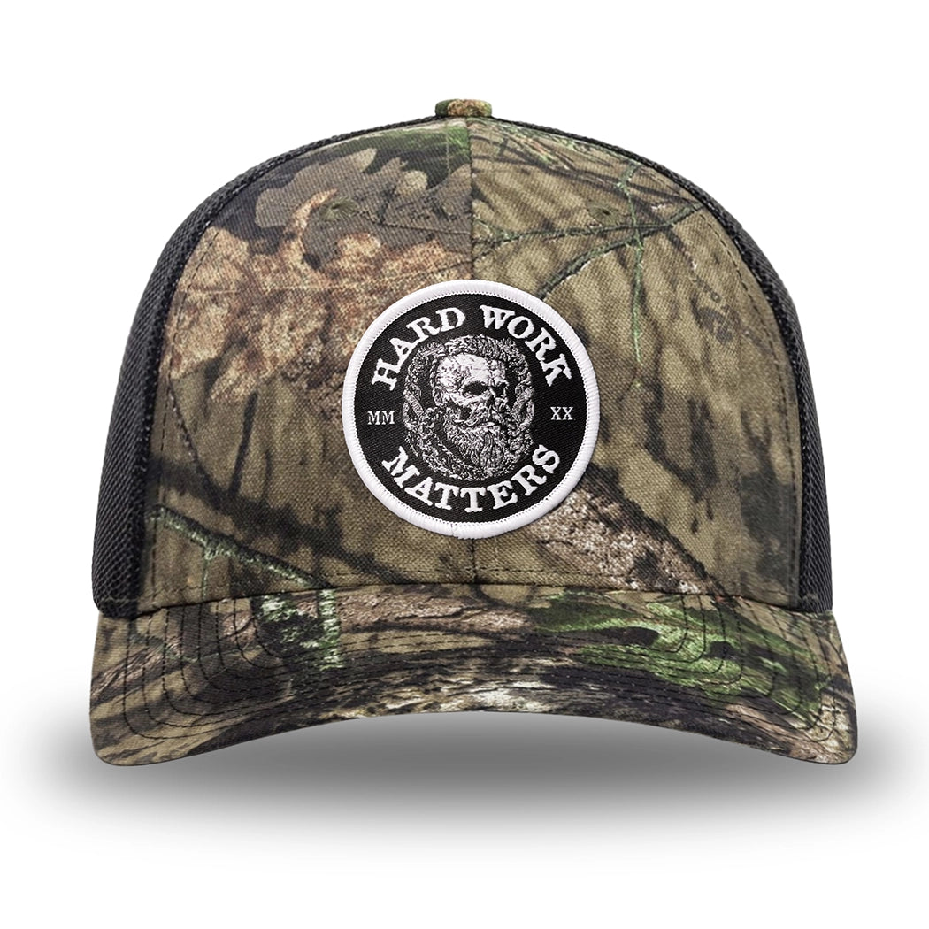 Mossy Oak/Country DNA/Black WeWorkin hat—Richardson 112 brand snapback, retro trucker classic hat style. HARD WORK MATTERS woven patch with white merrowed edge, on a black background with HARD WORK MATTERS text encircling a Viking-style skull center graphic with MM XX on the left and right respectively—patch is centered on the front panels.