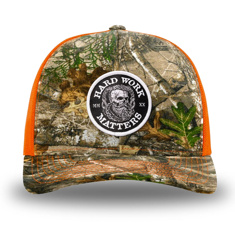 Neon Orange and RealTree Camo two-tone WeWorkin hat—Richardson 112 brand snapback, retro trucker classic hat style. HARD WORK MATTERS woven patch with white merrowed edge, on a black background with HARD WORK MATTERS text encircling a Viking-style skull center graphic with MM XX on the left and right respectively—patch is centered on the front panels.