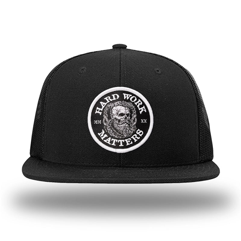 Solid Black WeWorkin hat—Richardson 511 brand snapback, flatbill trucker hat style. HARD WORK MATTERS woven patch with white merrowed edge, on a black background with HARD WORK MATTERS text encircling a Viking-style skull center graphic with MM XX on the left and right respectively—patch is centered on the front panels.