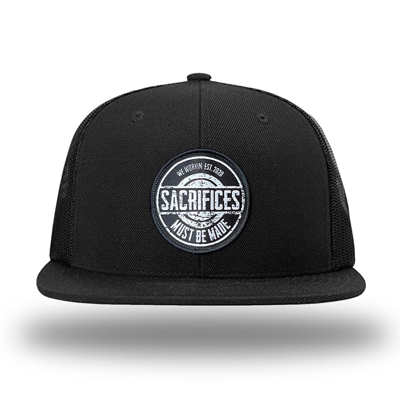 Solid Black WeWorkin hat—Richardson 511 brand snapback, flatbill trucker hat style. WeWorkin "SACRIFICES Must Be Made" circular woven patch, with black/white thread colors and black merrowed edge, is centered on the front panels.