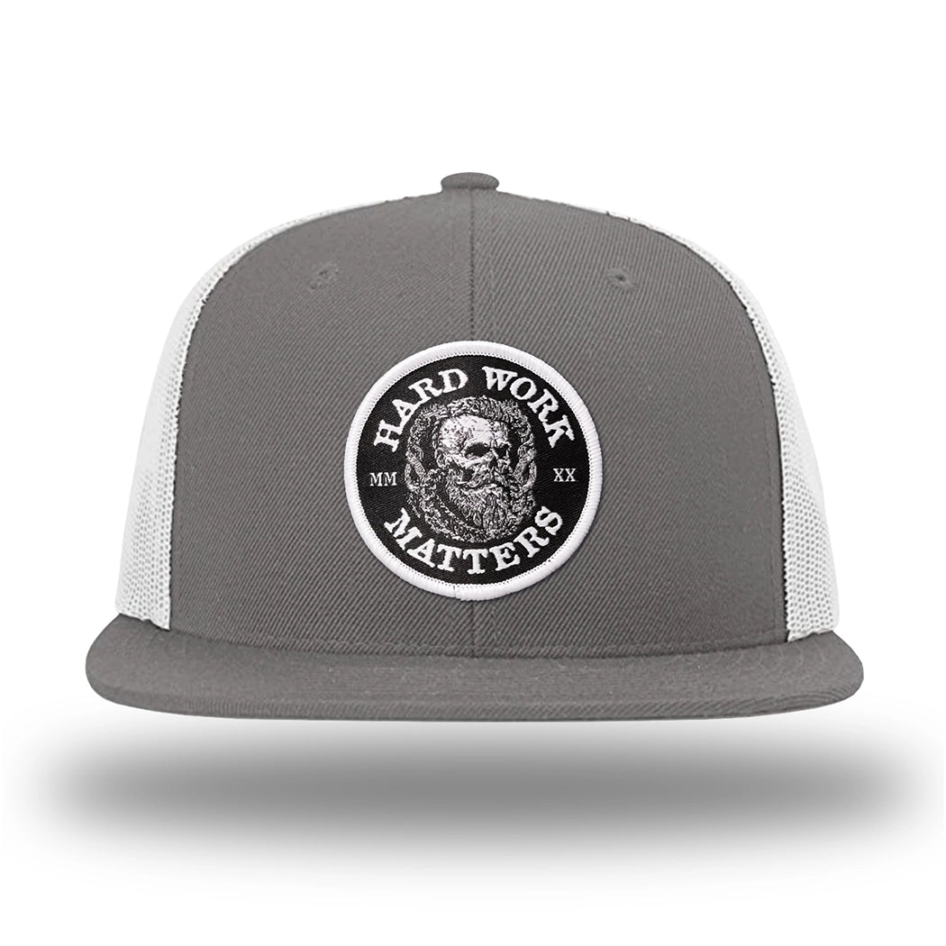 Charcoal/White WeWorkin hat—Richardson 511 brand snapback, flatbill trucker hat style. HARD WORK MATTERS woven patch with white merrowed edge, on a black background with HARD WORK MATTERS text encircling a Viking-style skull center graphic with MM XX on the left and right respectively—patch is centered on the front panels.