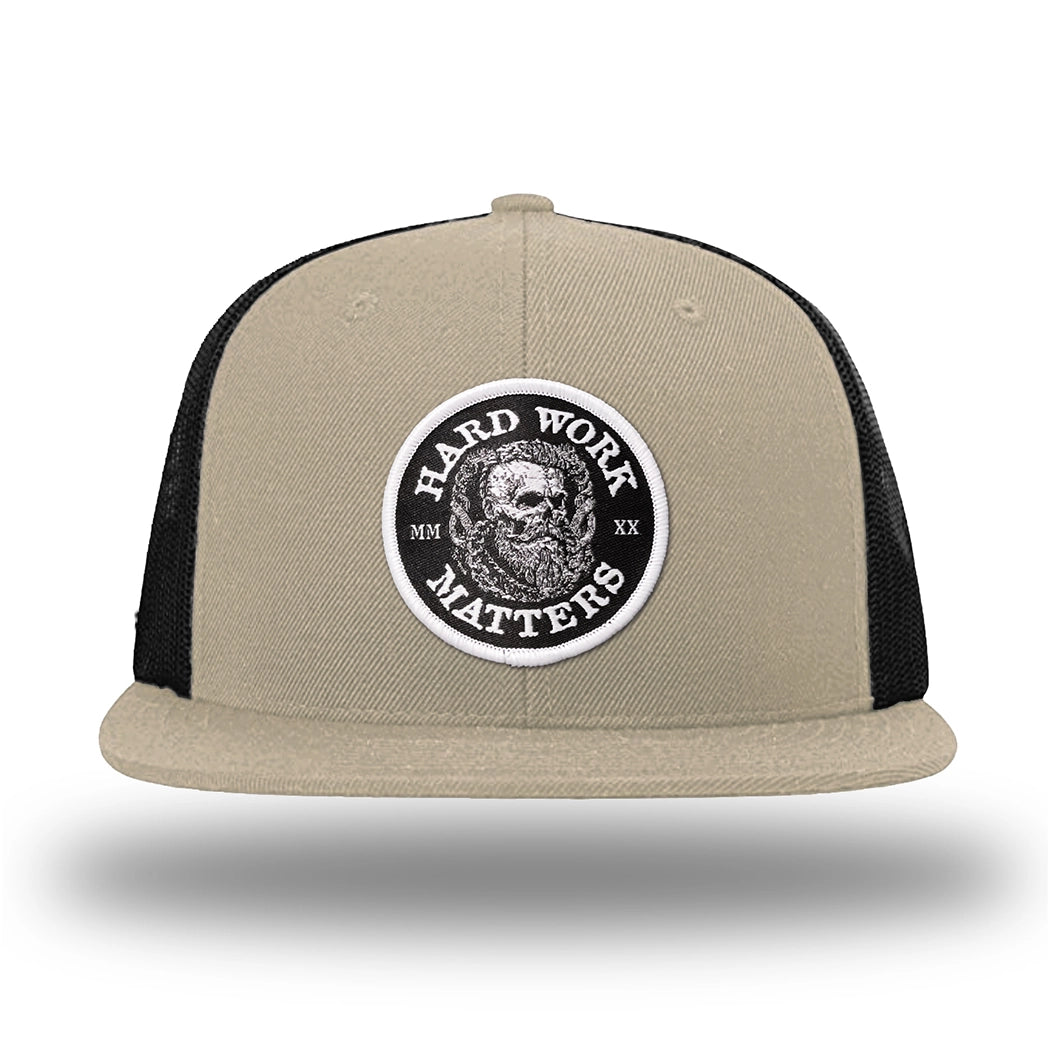 Khaki/Black WeWorkin hat—Richardson 511 brand snapback, flatbill trucker hat style. HARD WORK MATTERS woven patch with white merrowed edge, on a black background with HARD WORK MATTERS text encircling a Viking-style skull center graphic with MM XX on the left and right respectively—patch is centered on the front panels.